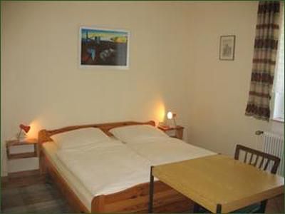 Ein einfaches Schlafzimmer mit einem Doppelbett und einem Tisch. An der Wand hängt ein Gemälde, und es gibt Tageslicht durch ein Fenster.