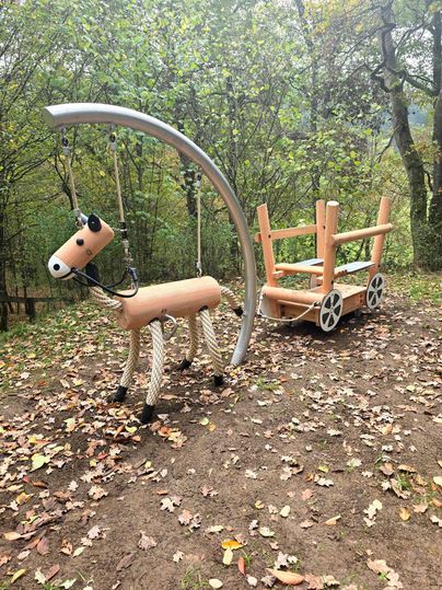 Een speelplaats met een houten paard en een koets. De omgeving is gekenmerkt door bomen en herfstbladeren.