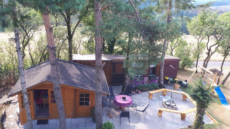 Ein gemütliches Ferienhaus umgeben von Bäumen. Davor befinden sich eine Terrasse mit Sitzgelegenheiten und ein Spielplatz.