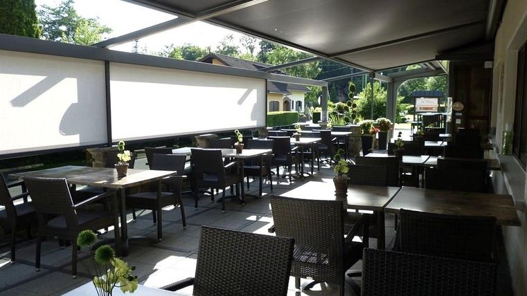 Ein gemütlicher Außenbereich eines Cafés mit mehreren Tischen und Stühlen. Die Terrasse ist mit Pflanzen dekoriert und hat einen klaren Himmel im Hintergrund.