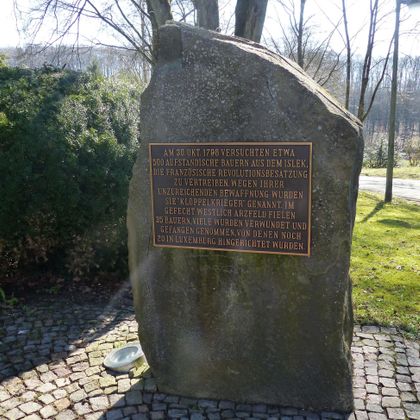 A large stone monument with an inscription in German. The stone commemorates a historical event and is located in a green environment.