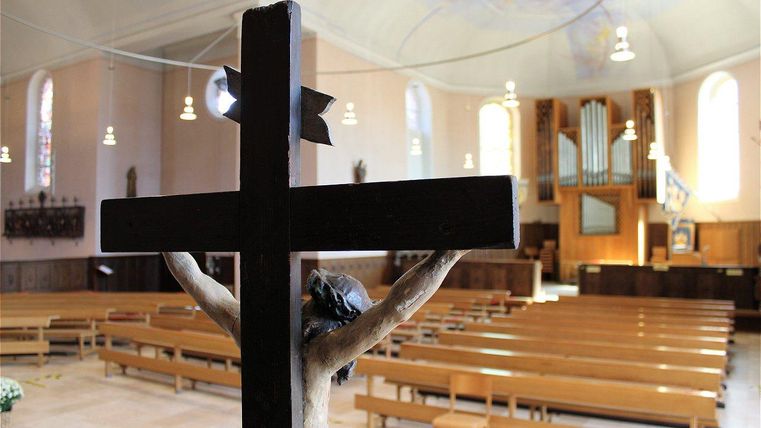 Eine stille Kirche mit einer Holzfigur am Kreuz im Vordergrund. Im Hintergrund sind Bänke und eine Orgel sichtbar.