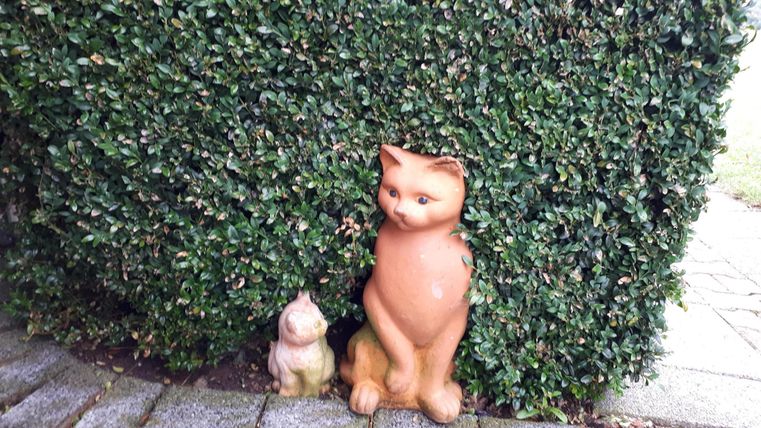 Zwei dekorative Katzenfiguren stehen neben einer Hecke. Die größere Figur ist braun und die kleinere ist hellfarben.