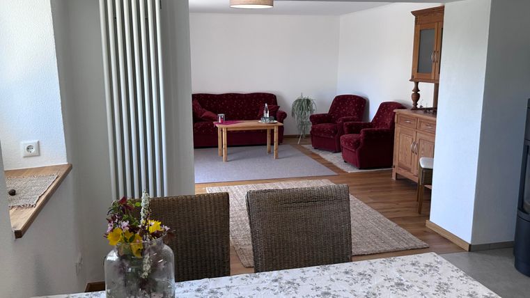 A cozy living room with red sofas and a wooden table. In the foreground, there is a table with a vase and fresh flowers.