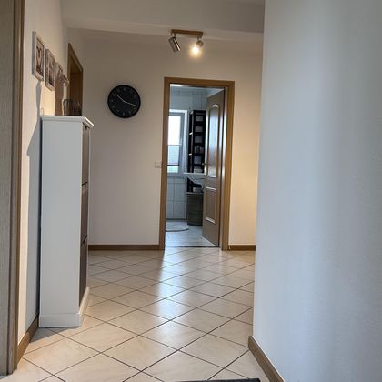 Ein einfacher Flur mit Fliesenboden. Auf der linken Seite steht ein weißer Schrank und im Hintergrund sieht man eine geöffnete Tür zu einem Raum.