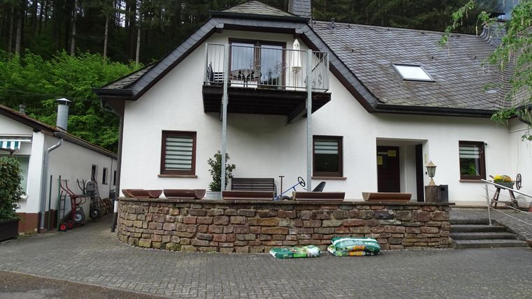 Ein zweigeschossiges Haus in ländlicher Umgebung mit einem Balkon. Im Hintergrund sind Bäume zu sehen, und der Außenbereich ist gut gepflegt.