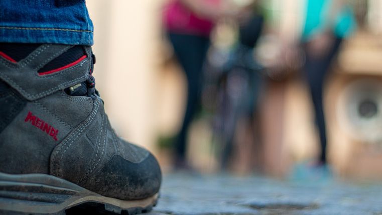Ein Wanderstiefel steht auf einem Pflasterweg. Im Hintergrund sind unscharfe Personen zu sehen, die sich unterhalten.