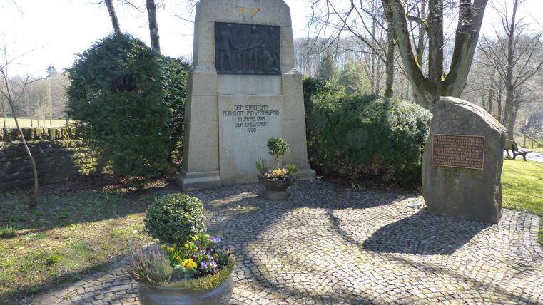 Ein historisches Gebäude mit Türmen steht vor einer bewaldeten Hügelkulisse. Der Platz zeigt eine ruhige Atmosphäre und ist von einigen Autos gesäumt.