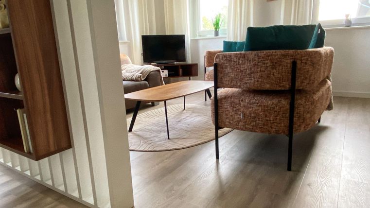 Een modern woonkamervoorziening met een comfortabele fauteuil en een houten salontafel. Grote ramen zorgen voor veel daglicht en een aangename sfeer.