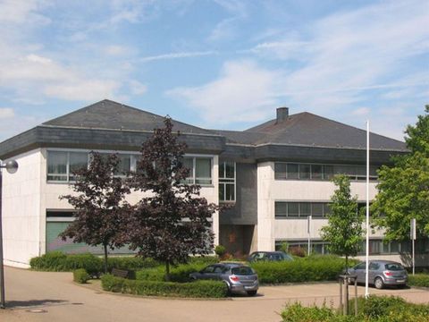 Ein modernes Gebäude mit einem Steildach und großen Fenstern. Umgeben von Bäumen und einigen geparkten Autos.