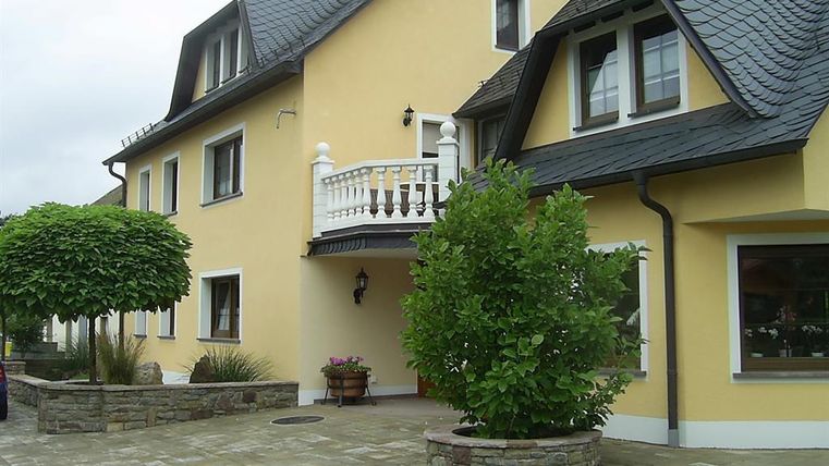 Une maison moderne avec une façade jaune et un balcon. Devant la maison, il y a des arbres et une surface en pavés.