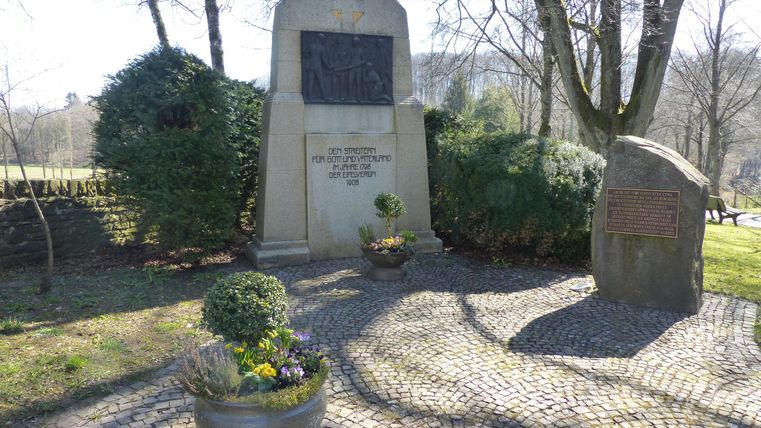 A monument in a quiet setting with trees and shrubs. The area is paved with gravel and contains flower pots.