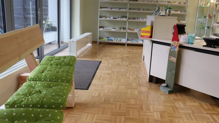 Ein heller Raum mit Holzboden und großen Fenstern. Auf der linken Seite befindet sich eine grüne Bank und ein Empfangstresen auf der rechten Seite.