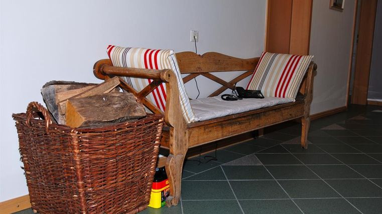 Un banc en bois confortable avec des coussins se trouve dans un couloir. À côté, il y a un panier avec des morceaux de bois.