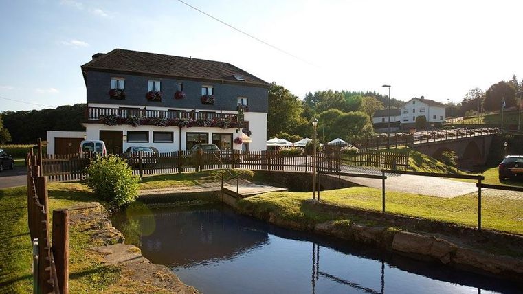 Ein gemütliches Haus mit Balkonen und einem ruhigen Teich davor. Im Hintergrund sind grüne Wiesen und Bäume zu sehen.