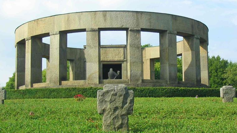 Ein runder, steinerner Bau mit offenen Fensteröffnungen steht inmitten von Grün. Im Vordergrund sind einige grasbewachsene Grabsteine zu sehen.