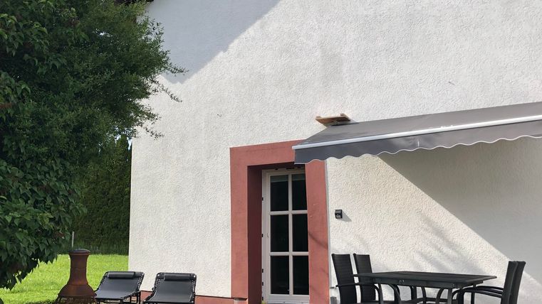 Ein modernes Haus mit einer weißen Fassade und roter Umrandung. Auf der Terrasse stehen ein Tisch und Stühle sowie Liegen im Garten.