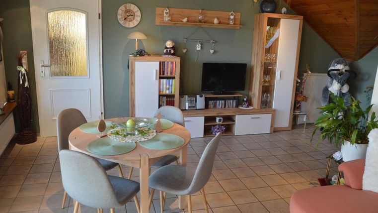 Ein gemütlicher Wohnraum mit einem runden Tisch und modernen Stühlen. An den Wänden sind Bücherregale und eine TV-Ecke zu sehen.