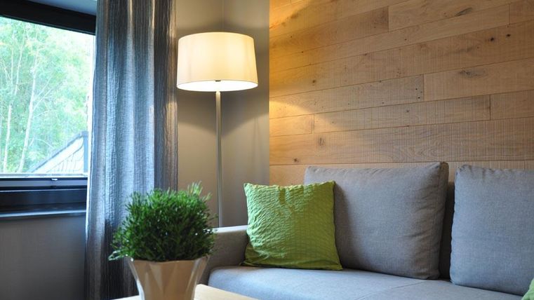 Ein gemütlicher Wohnbereich mit einem grauen Sofa und grünen Kissen. Eine Stehlampe und ein Fenster mit Vorhängen ergänzen das moderne Ambiente.