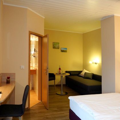 Ein gemütliches Hotelzimmer mit einem Doppelbett, einem Tisch, einem Stuhl und einer Sitzecke. Die Wände sind in sanften Farben gehalten, und ein Fernseher ist an der Wand montiert.