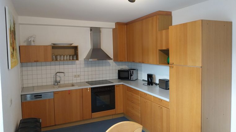 Eine moderne Küche mit holzfarbenen Schränken und weißen Fliesen. Sie ist ausgestattet mit einem Herd, einem Kühlschrank und verschiedenen Küchengeräten.