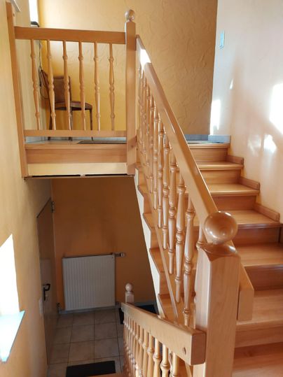 Een heldere trap met een houten leuning leidt naar boven. Op de achtergrond is een stoel en een muur in warme kleuren te zien.