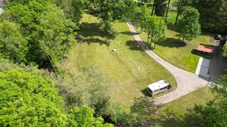 Eine grüne Wiese umgeben von Bäumen, mit einem kleinen Weg, der durch die Landschaft führt. Im Hintergrund steht ein Wohnmobil und eine Picknickbank.