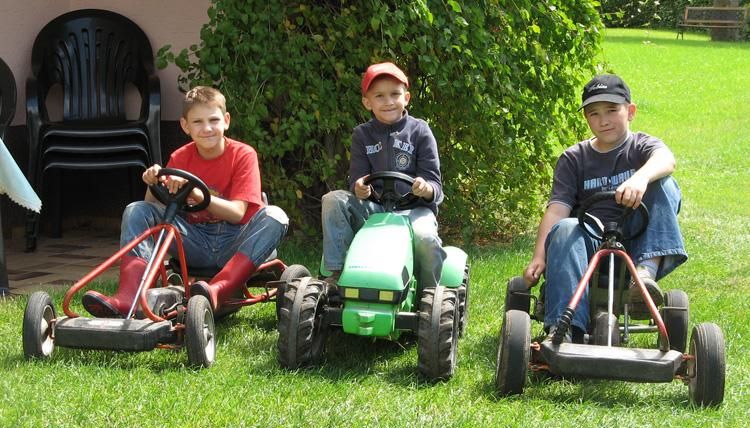 Drei Kinder sitzen auf Kettcars auf einer grünen Wiese. Sie lächeln und haben Spaß miteinander.