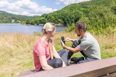 Ein Paar sitzt auf einer Bank am Ufer eines Gewässers. Sie schauen auf ein Smartphone und genießen die schöne Natur um sich herum.