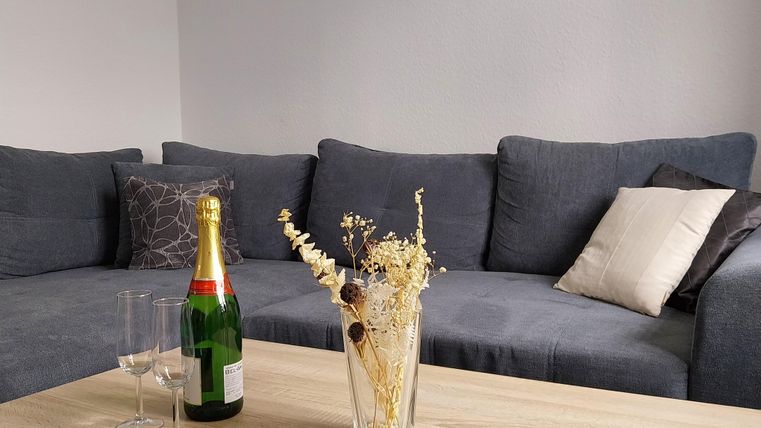 Ein gemütliches Wohnzimmer mit einer grauen Couch und einem Holztisch. Auf dem Tisch stehen eine Flasche Sekt, Gläser und eine Vase mit Trockenblumen.
