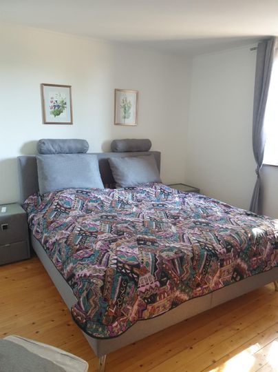 Ein gemütliches Schlafzimmer mit einem großen Bett und buntem Bettzeug. An den Wänden hängen zwei Bilder und ein Fenster sorgt für viel Licht.