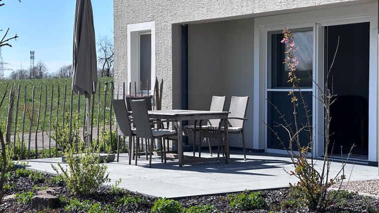 A cozy seating area on a terrace with a table and chairs. In the background, green meadows and a sun umbrella are visible.