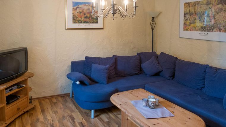 Ein gemütliches Wohnzimmer mit einem blauen Sofa und einem Holzkaffeetisch. Im Hintergrund gibt es einen Fernseher und Bilder an der Wand.
