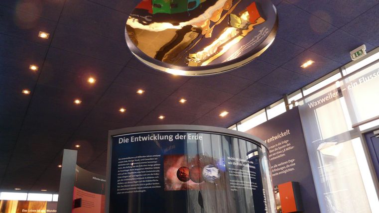 Ein moderner Ausstellungsraum mit interaktiven Tafeln zur Erdgeschichte. Die Decke reflektiert Licht und zeigt ein faszinierendes Design.