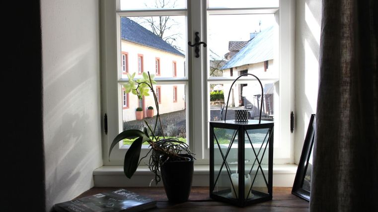 Ein heller Fensterausschnitt mit Blick auf ein ruhiges Dorf. Auf der Fensterbank stehen eine Pflanze und eine Laterne.