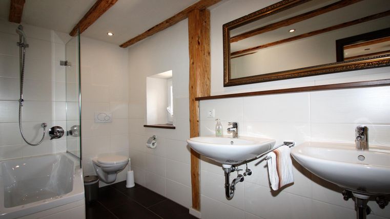 Ein modernes Badezimmer mit hellen Fliesen und Holzakzenten. Es gibt eine Badewanne, zwei Waschbecken und einen großen Spiegel.