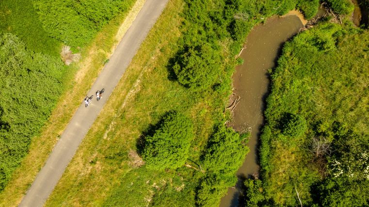 Een groene landschap met een smal pad en een kleine rivier. Enkele personen zijn langs het pad onderweg.