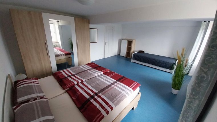 Ein modernes Schlafzimmer mit einem großen Doppelbett und einem Einzelbett. Der Raum ist hell und freundlich, mit einem blauen Fußboden und minimaler Einrichtung.