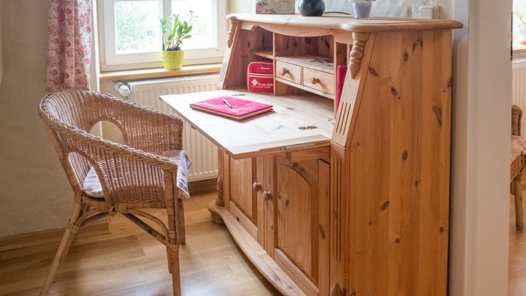 Ein Holzschreibtisch mit einem Stuhl aus Rattan steht in einem hellen Raum. Auf dem Tisch liegt ein rotes Notizbuch und vor dem Fenster steht eine Pflanze.