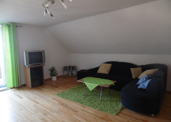 Ein gemütliches Wohnzimmer mit einem dunkelblauen Sofa und einem kleinen Tisch. Ein Fernseher steht in der Ecke, und grüne Vorhänge lassen viel Licht herein.