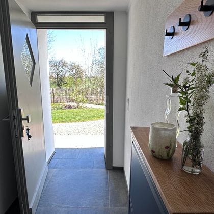 A bright entrance area with an open door and a view of the outdoors. On a wooden shelf, there are vases with flowers.