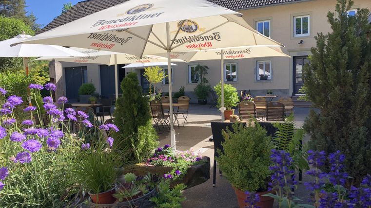 Ein gemütlicher Außenbereich mit weißen Sonnenschirmen und blühenden Pflanzen. Tische und Stühle laden zum Verweilen ein.