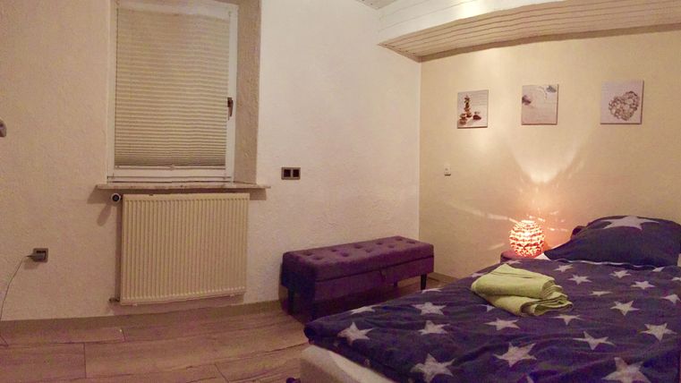 Une chambre confortable avec un lit, un banc et une fenêtre. Des images sont accrochées au mur et une lumière douce crée une atmosphère agréable.