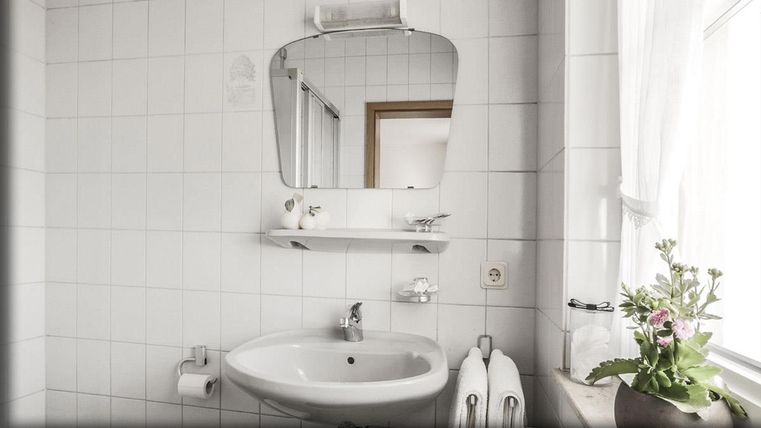 Ein helles Badezimmer mit weißen Fliesen und einem runden Spiegel über dem Waschbecken. Auf dem Regal sind frische Blumen und Handtücher sichtbar.