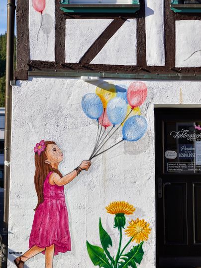 Ausschnitte einer weißen Hauswand darauf ist ein Kind mit bunten Luftballons gemalt. 