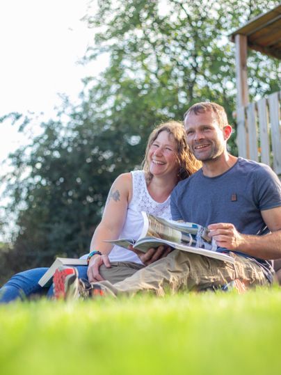 Ein lächelndes Paar sitzt auf dem Rasen und schaut in ein Magazin. Im Hintergrund sind Bäume und eine Holzstruktur sichtbar.
