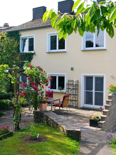 Un jardin accueillant avec une terrasse et des buissons de fleurs. À l'arrière-plan, il y a une maison lumineuse avec de grandes fenêtres.