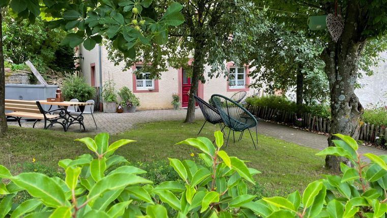 A cozy garden with trees and a terrace. In the background, there is a bright house with red windows.