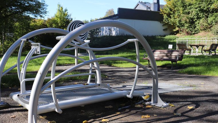 Ein modernes Fitnessgerät im Freien in einem Park. Im Hintergrund sind Bäume und Sitzgelegenheiten sichtbar.