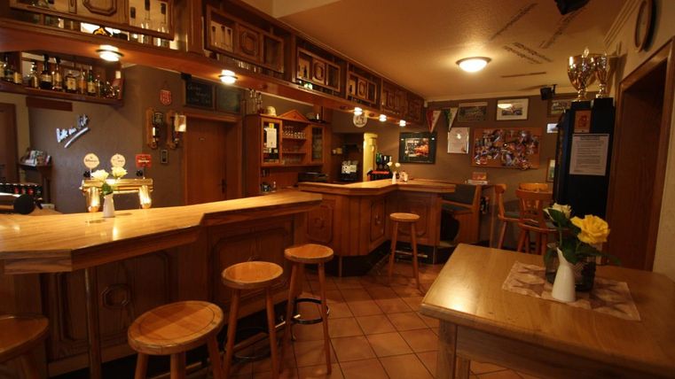 Eine gemütliche Bar mit Holztheke und Hockern. An den Wänden hängen Bilder und es gibt eine freundliche Atmosphäre.